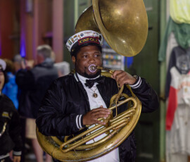 Musician in Kinfolk hat and black jacket plays sousaphone on a lively city street at night, evoking Louisiana wedding vibes.