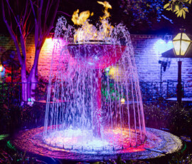 A round fountain with colorful lights, water jets, and flames at night—ideal for a Louisiana wedding with brick wall and trees.