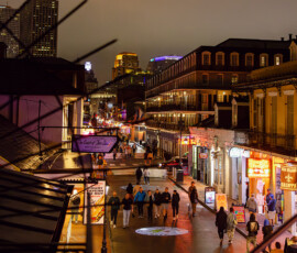 Lively city street at night with people, neon signs, lit buildings, wet pavement reflections, and a cloudy sky.