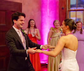 Smiling couple dances at a wedding as guests in colorful dresses watch under pink lights, perfect Gulf South wedding inspiration.