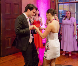 A suited man and barefoot bride dance as three women watch and smile in a warmly lit, pink Gulf South wedding reception.
