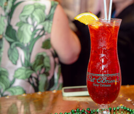 Red cocktail with orange slice, straws, and Mardi Gras beads at Pat O’Brien’s New Orleans; festive guests blurred behind.