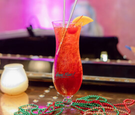 Red cocktail with orange garnish and straws on a piano with Mardi Gras beads, candle, and pink background—festive wedding vibe.