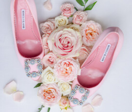 Manolo Blahnik pink jeweled ballet flats on white, surrounded by pink and white roses, green leaves, and rose petals.