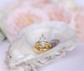 Gold wedding band and marquise diamond ring on oyster shell with soft pink and white flowers blurred in the background.