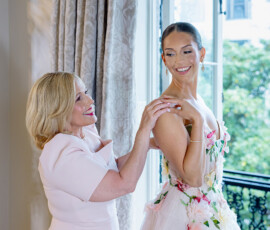 Two women, one in a pale dress and one in a floral gown, smile by a window before a wedding; greenery is visible outside.