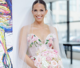 Bride in floral wedding dress and veil holds pink and white roses indoors by colorful painting, joyful before New Orleans ceremony.