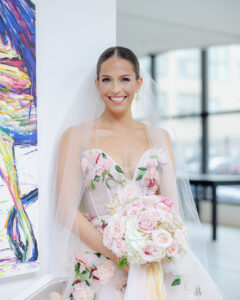 Bride wearing a floral-embroidered wedding gown and veil holds a soft pink rose bouquet while smiling indoors near a window.