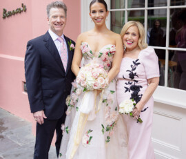 Bride in floral dress smiles between two adults with bouquets outside a pink building at a New Orleans wedding.