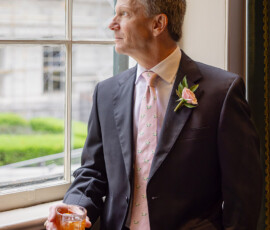 A suited man with a pink tie and boutonniere holds a drink, gazing out a window at greenery—elegant wedding inspiration.