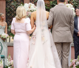 Bride in floral gown stands between two adults at a flower-adorned outdoor New Orleans wedding altar, seen from behind.