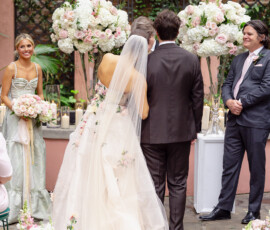 Bride in floral wedding dress stands with groom at outdoor Louisiana ceremony, with flowers and guests watching the vows.