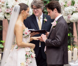 Bride and groom exchange rings outdoors, surrounded by white and pink flowers and candles at their wedding ceremony.