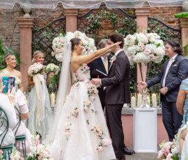 Bride in floral dress smiles, touching groom’s face at outdoor Gulf South wedding with pink and white flowers.