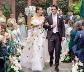 Bride in floral dress and groom in dark suit walk down a flower-lined aisle, smiling at a Louisiana wedding ceremony.