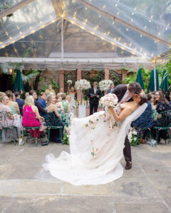Groom dips the bride for a kiss during an outdoor wedding ceremony under a clear tent as guests look on.