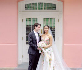 Bride in floral gown and groom in suit smile outside pink building 417 after Gulf South wedding ceremony.