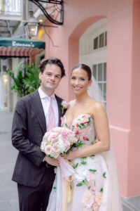 Bride in a floral-embroidered wedding gown and veil stands with the groom holding a pink and white rose bouquet on a pastel-colored street.