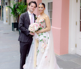 Smiling bride in floral gown and groom in dark suit on a sidewalk by a pink building at their joyful New Orleans wedding.