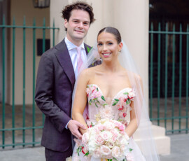 Bride in floral dress with rose bouquet and groom in dark suit smile outdoors by classic New Orleans building and iron fence.