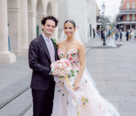 Bride in floral strapless gown and veil with groom in dark suit pose, smiling on a city street outside a historic building.
