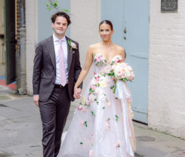 Smiling couple walks hand in hand—he in a dark suit, she in a floral gown—by a white brick building on their wedding day.