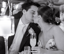 A formally dressed couple kisses at a decorated table with champagne, elegant flowers, and soft lighting—a romantic wedding scene.