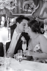 Black-and-white wedding reception moment of the bride and groom sharing a kiss at their candlelit table, holding champagne flutes during an elegant New Orleans celebration.