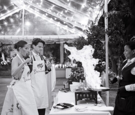 Couple in aprons watches chef cook with dramatic flame under string lights at a lively outdoor New Orleans wedding event.