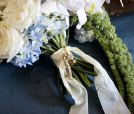 Bridal bouquet featuring white roses, orchids, and soft blue hydrangeas, wrapped with flowing ivory ribbon and a vintage brooch detail.