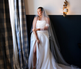 Bride stands by a window in a flowing white wedding gown with a long veil, softly lit by natural light.
