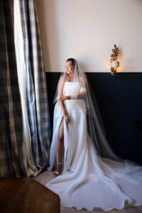 Bride stands by a window in a flowing white wedding gown with a long veil, softly lit by natural light.