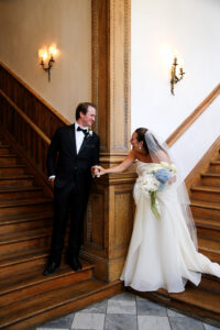 Bride and groom hold hands and smile at each other on a wooden staircase, with the bride holding a white and blue bouquet.