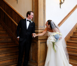 Bride and groom hold hands and smile at each other on a wooden staircase, with the bride holding a white and blue bouquet.