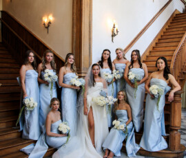 Bride stands on a grand staircase surrounded by bridesmaids in pale blue dresses, each holding white and blue floral bouquets.