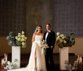 Bride and groom posing together at the altar, featuring modern floral arrangements, candlelight, and a textured wall backdrop in an elegant indoor wedding setting.