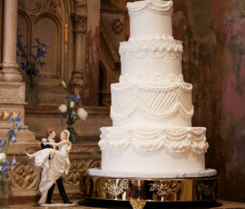 Classic tiered white wedding cake with ornate piping details and a bride and groom cake topper, displayed on an elegant stand.