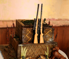 Groom’s cake designed with a rustic hunting theme, featuring sculpted rifles, ducks, cattails, and a dog detail.