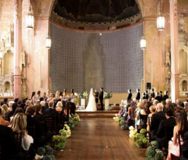 Wide view of a wedding ceremony inside a historic church, with guests seated along a flower-lined aisle as the couple stands at the altar.