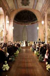 Wide view of a wedding ceremony inside a historic church, with guests seated along a flower-lined aisle as the couple stands at the altar.