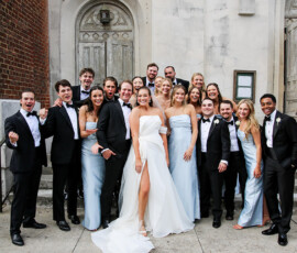 Bride and groom pose with their wedding party outside a church, surrounded by smiling bridesmaids in pale blue dresses and groomsmen in black tuxedos.