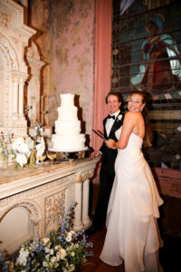 Bride and groom smile while cutting their wedding cake inside a historic venue, surrounded by white and blue floral details.
