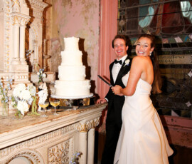 Bride and groom smile while cutting their wedding cake inside a historic venue, surrounded by white and blue floral details.