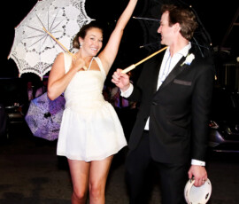 Bride and groom celebrate during a nighttime second line, smiling and dancing with umbrellas and tambourines.