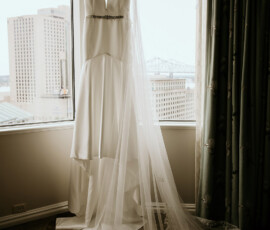 White wedding dress with long lace veil hangs by a window, city and bridge visible on a cloudy Louisiana day.