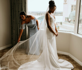 Bride in white gown by window as woman in blue adjusts lace train; natural light fills Louisiana wedding room.
