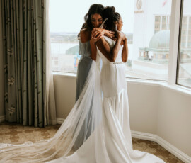 Two women embrace by a large window, one in a white lace wedding dress and one in gray, with city buildings outside.