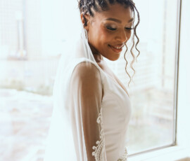 Bride in lace-sleeved white dress and veil smiles by window, city buildings behind her, ready for New Orleans wedding.