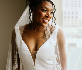 Smiling bride in white dress and veil by a window at her New Orleans wedding, wearing earrings, belt, and chest tattoo.