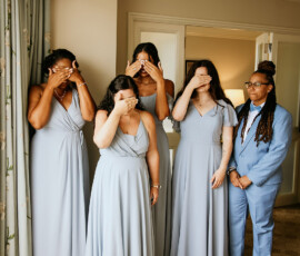 Five people pose in a room: four in matching blue dresses cover their faces, while one in a blue suit smiles at them.